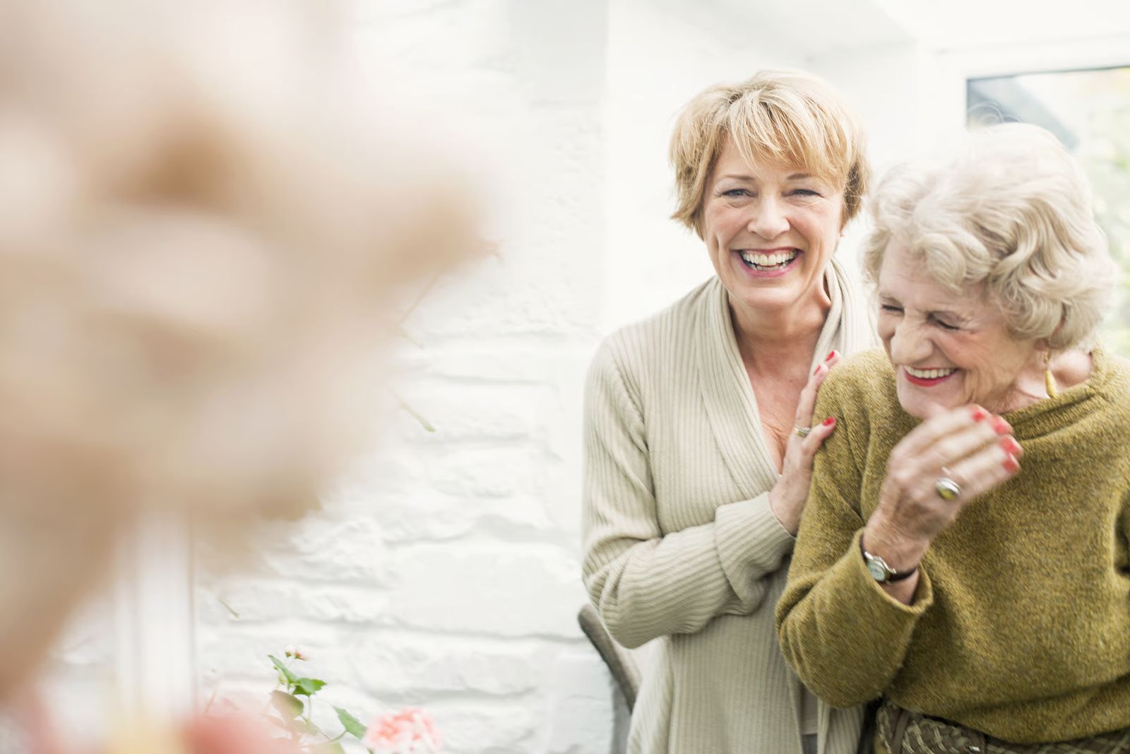Zwei Frauen lachen gemeinsam in einem hellen Wintergarten, die jüngere legt der älteren die Hand auf die Schulter